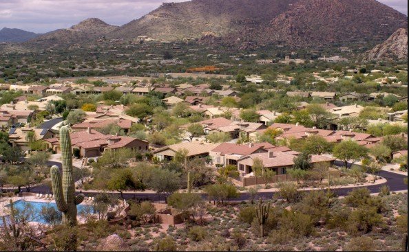 A desert neighborhood with houses, cacti, and a swimming pool, set against mountains in the background.