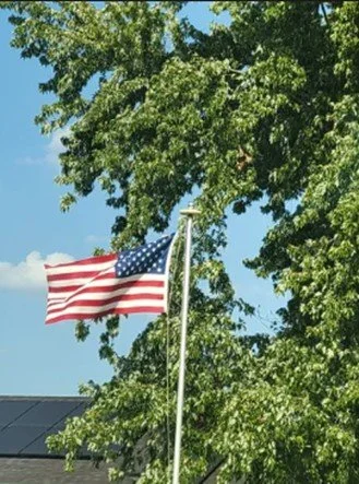 American flag flying outdoors among green trees and a blue sky.