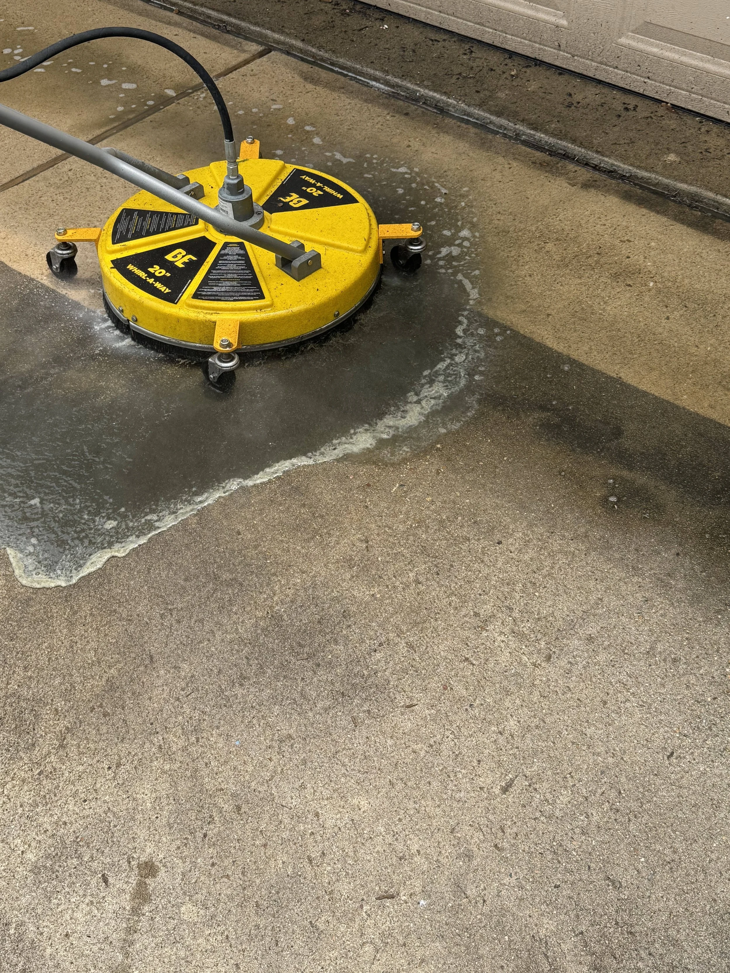 A disc cleaning machine on a concrete sidewalk, with soap and water around it, near a building with wooden siding.