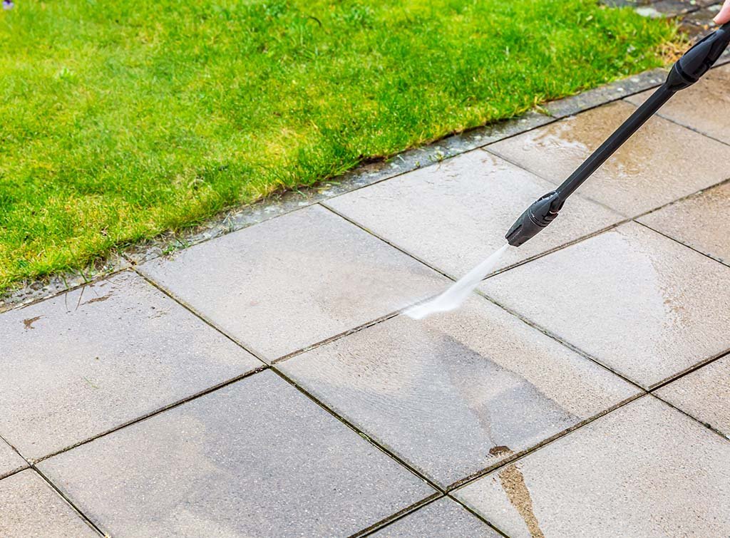 A person cleaning tiled outdoor walkway with a pressure washer, water spraying on the tiles, with a patch of green grass nearby.