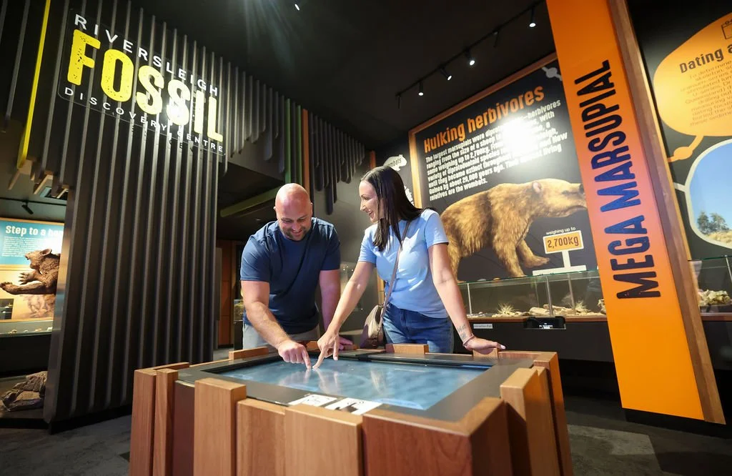A man and woman exploring a fossil exhibit at a museum's discovery center. They are pointing at a touch screen table. The background has informational signs about extinct species and a large bear.