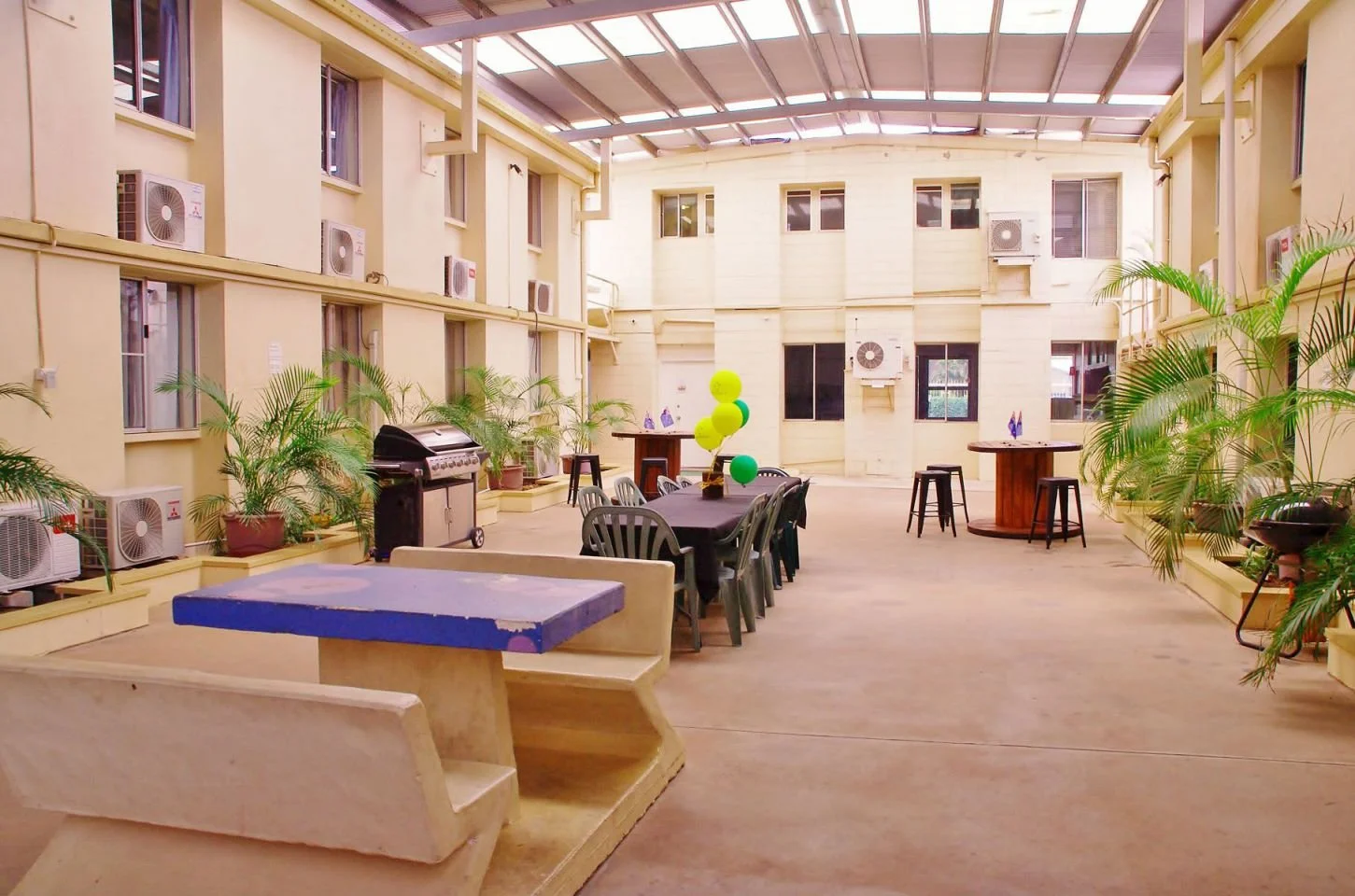 An outdoor patio area inside a courtyard of a building with yellow walls, multiple windows, and air conditioning units. There are potted plants, tables with chairs, balloons, and a barbecue grill.