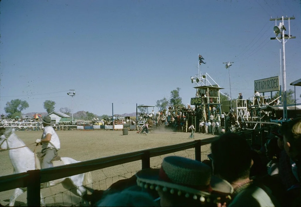 Historical image of Mount Isa Rodeo arena with a rider on a white horse in the foreground, spectators in the stands, and a ride or show going on in the background with people and structures on a sunny day.