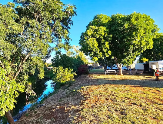View of a park with a small river on the left, several large green trees, a grassy area, and a recreational area with benches and trailers in the background.