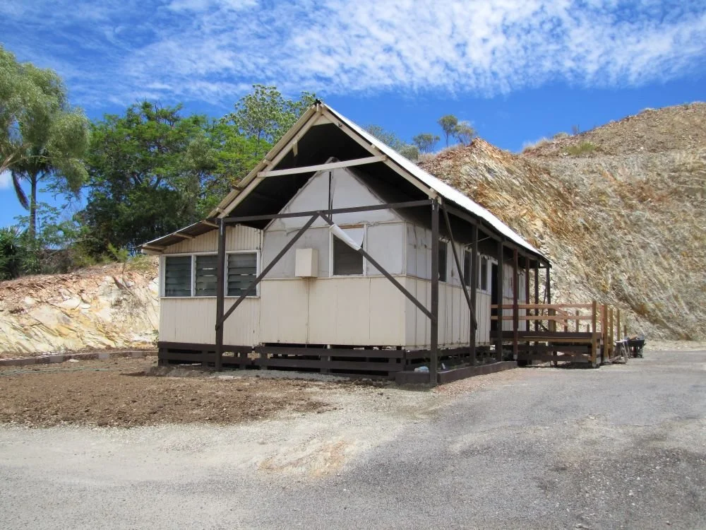 A Tent House with a white exterior, situated on a gravel lot with a dirt hillside behind it, and surrounded by green trees and a bright blue sky.