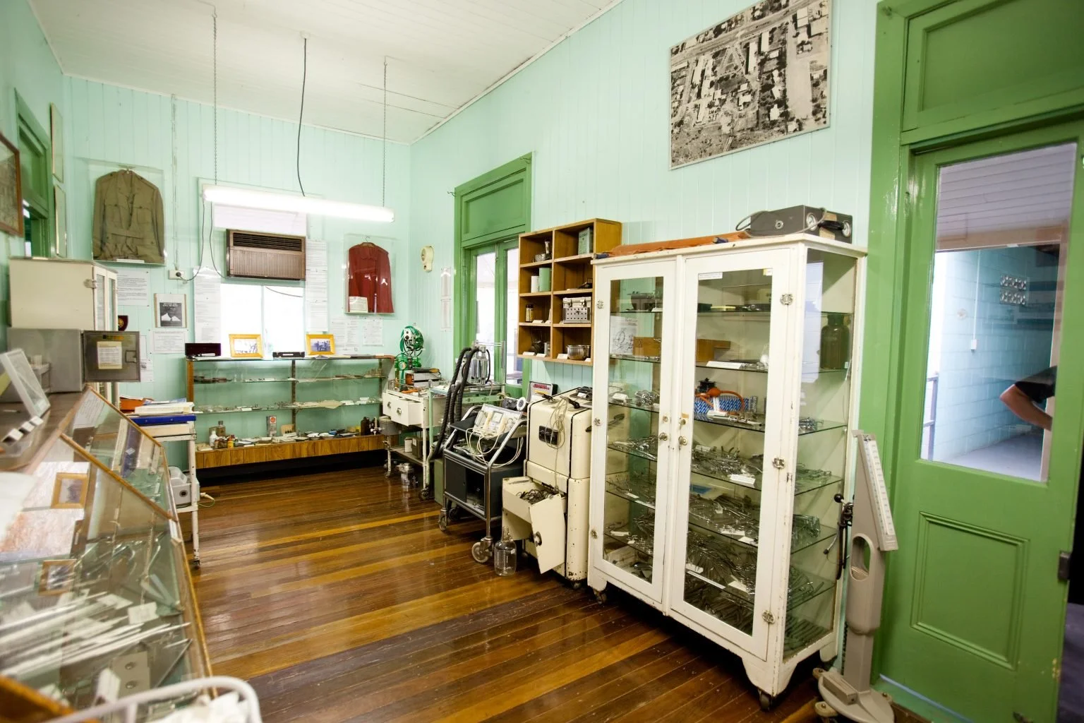 View of the Underground Hospital Museum with green walls, wooden floor, and glass display cabinets filled with small artifacts. There are various vintage medical or scientific equipment, framed pictures on the walls, and old national flags.