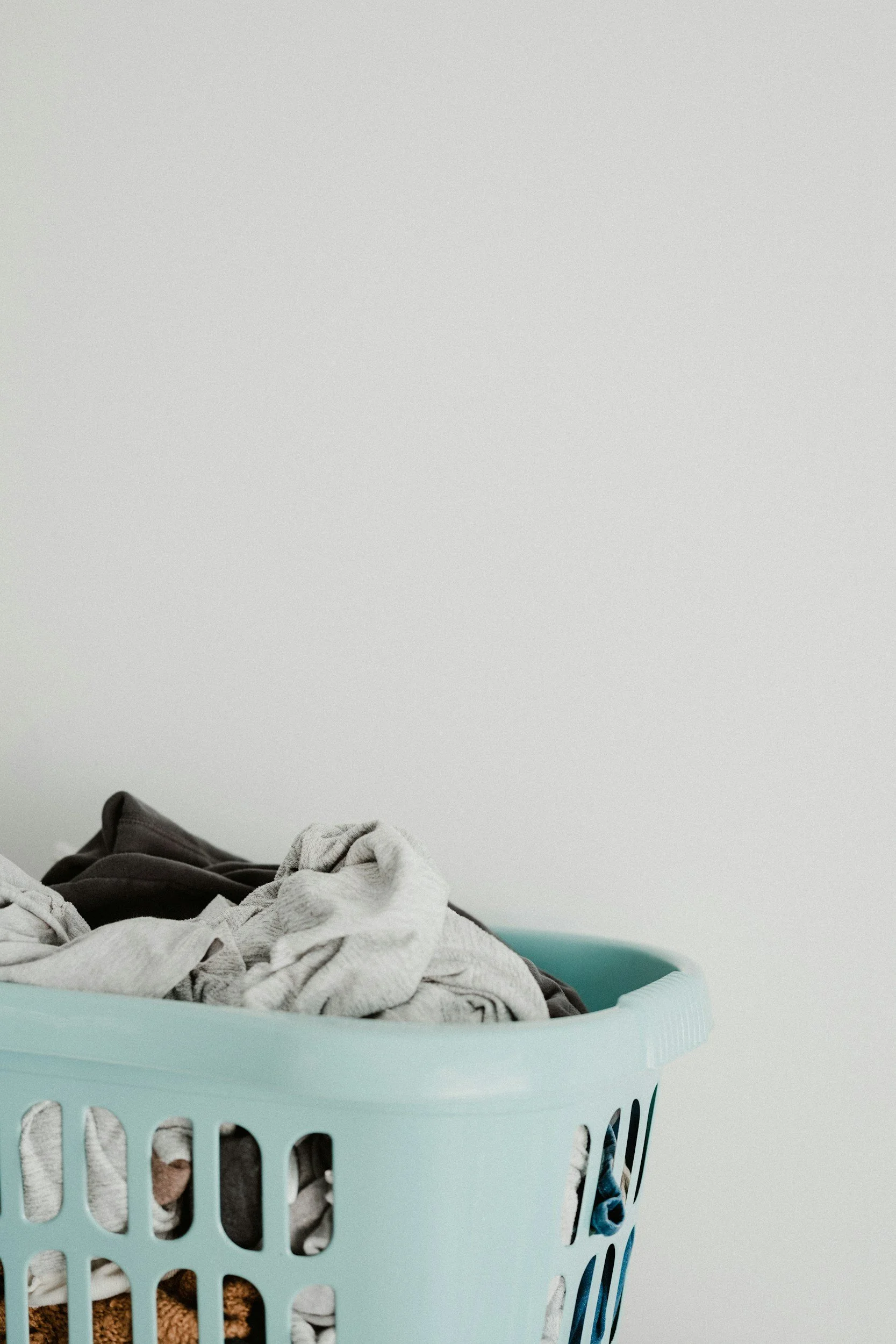 Clothes piled in a laundry basket with a white and black shirt visible at the bottom, against a plain white wall background.