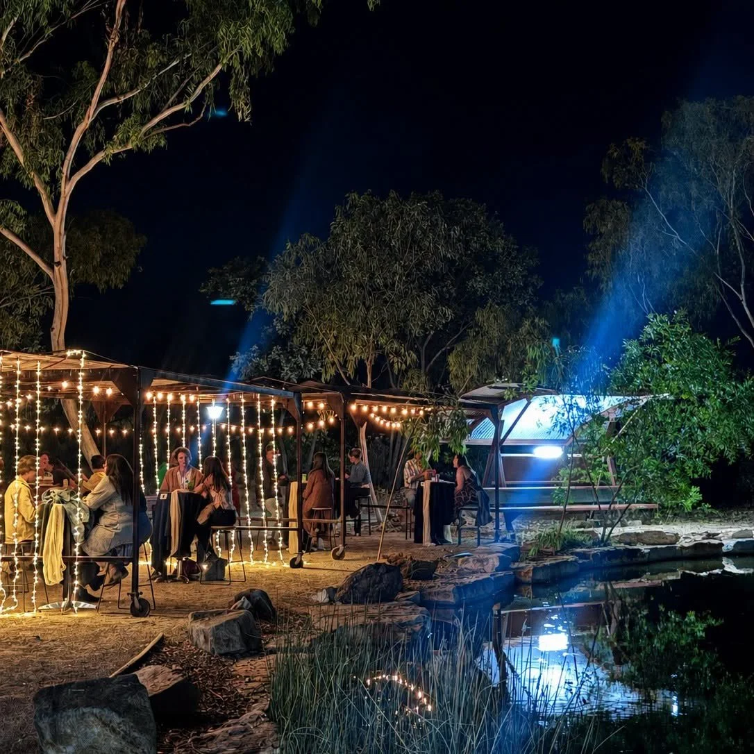 People dining at the Outback park decorated with string lights, near a body of water at night, surrounded by trees.