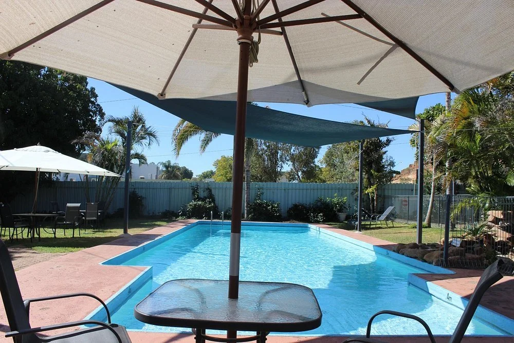 View of a backyard swimming pool with patio chairs and umbrellas, surrounded by lush trees and a blue fence.