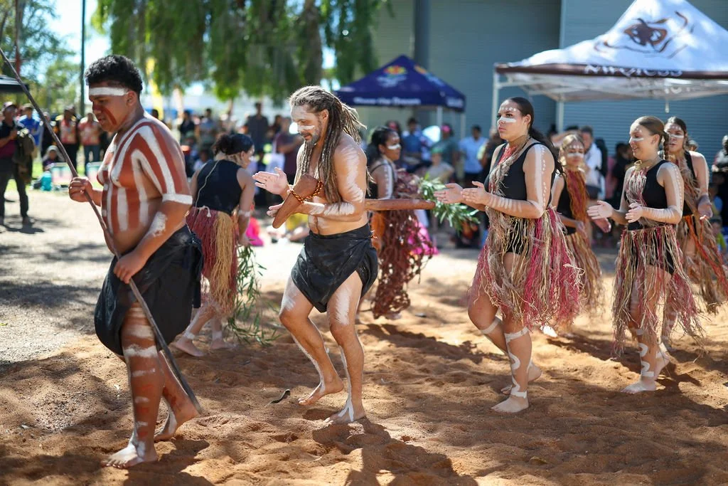 Indigenous people participating in a traditional dance or ceremony outdoors, dressed in traditional attire with body paint and grass skirts, with onlookers in background.