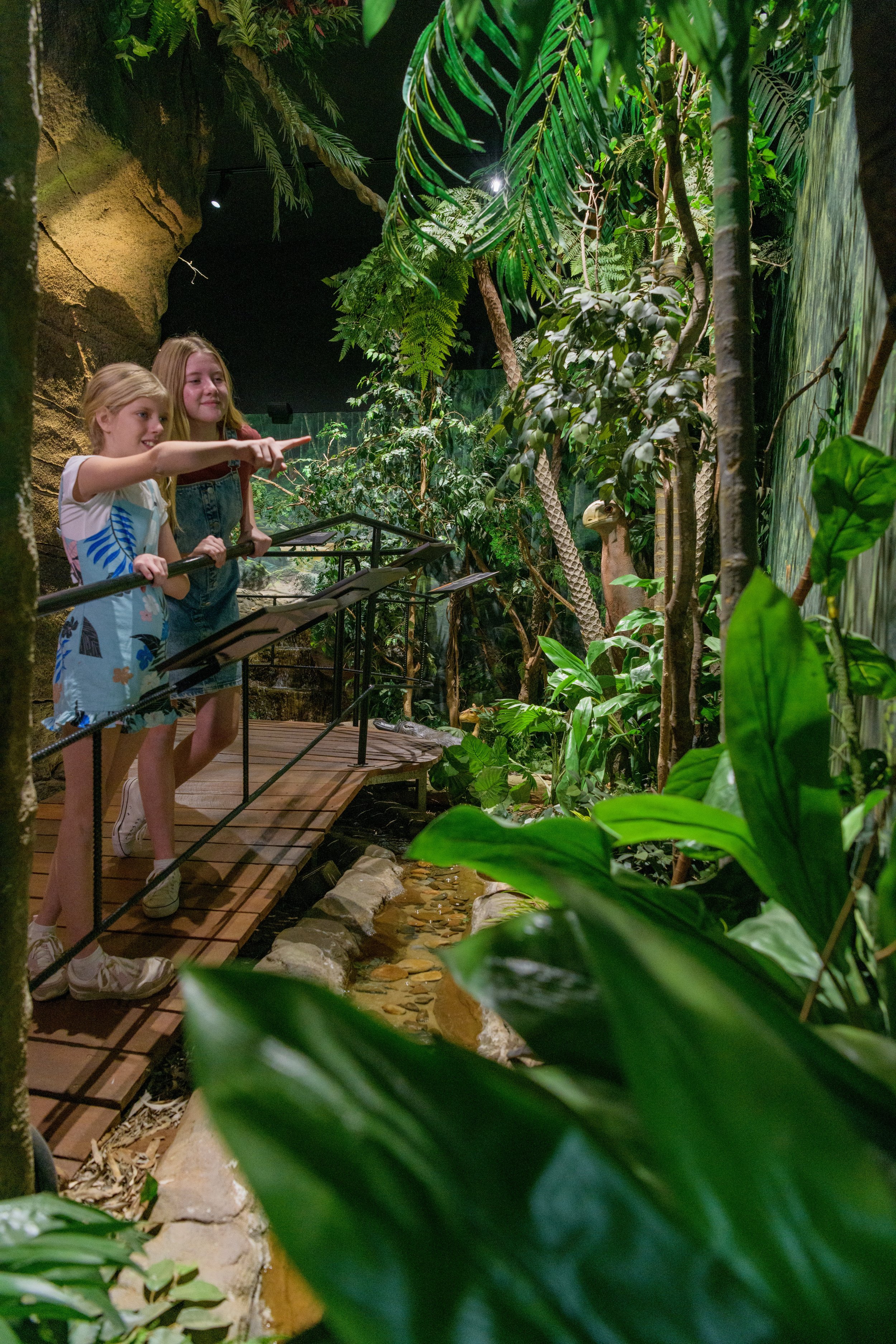 Two young girls are observing a small water feature in a lush, indoor tropical rainforest exhibit at the Riversleigh Discovery Centre. The setting is filled with tall trees and broad-leafed plants, creating a dense jungle atmosphere.