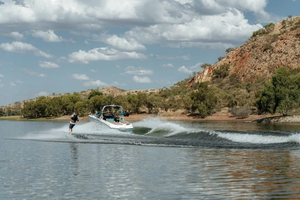 A person water skiing on a lake, with a motorboat towing them, surrounded by hills and trees under a partly cloudy sky.