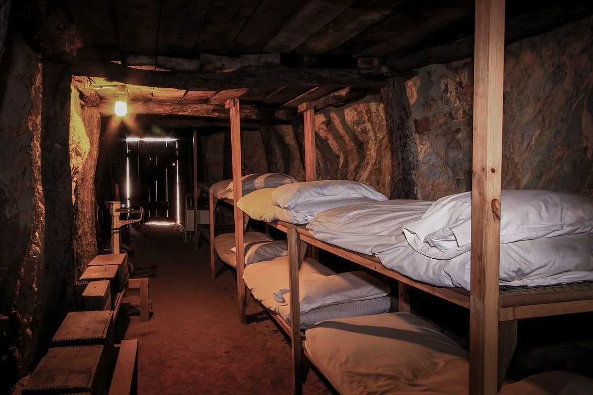 Underground Hospital Museum with wooden bunks and bedding, illuminated by a dim light and a small opening at the end of the passage.