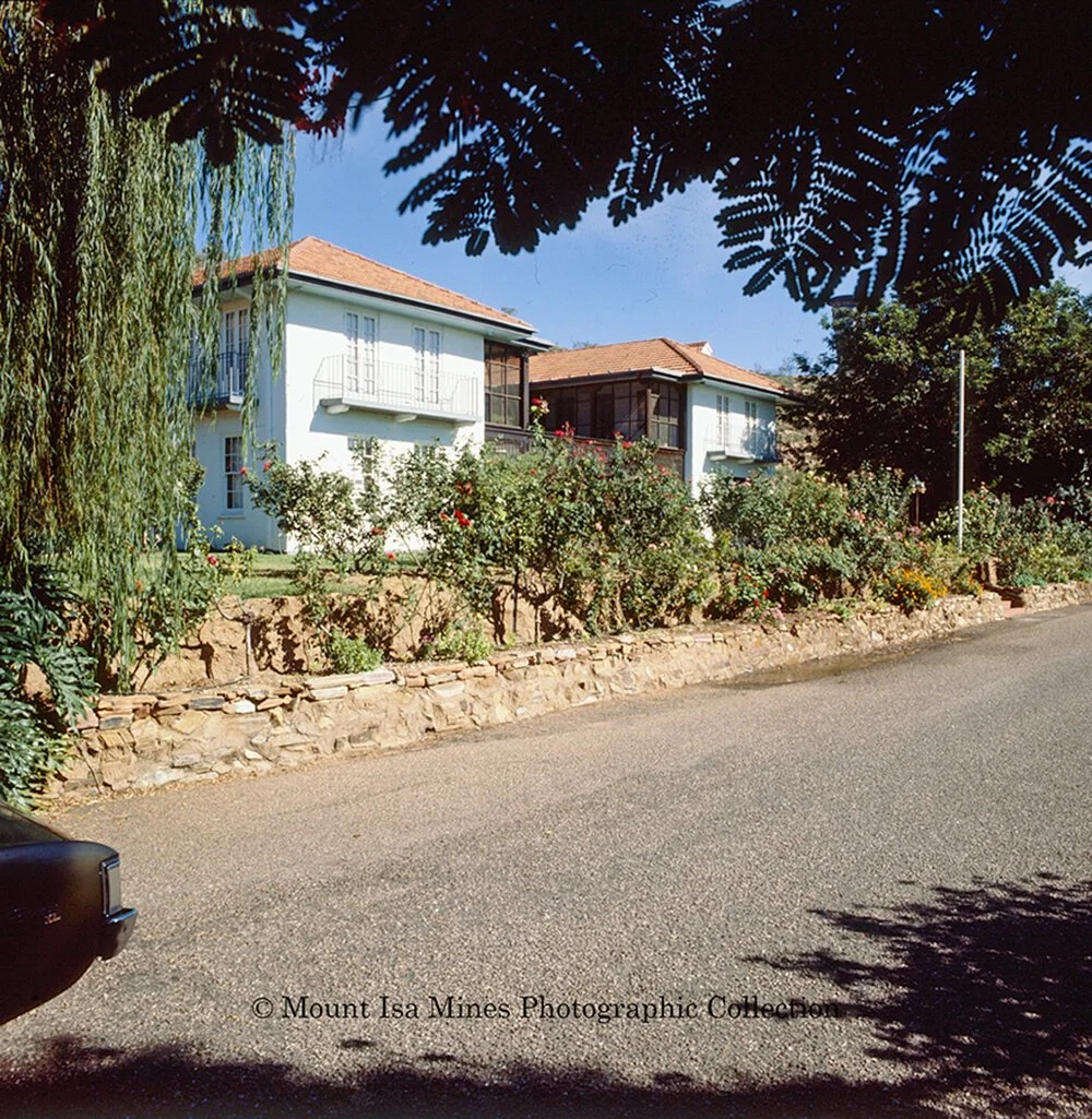A residential street with a white two-story house, surrounded by a garden with assorted plants and flowers. A stone retaining wall lines the garden, and a black car is partially visible in the lower left corner. Shadows from trees are cast on the street.