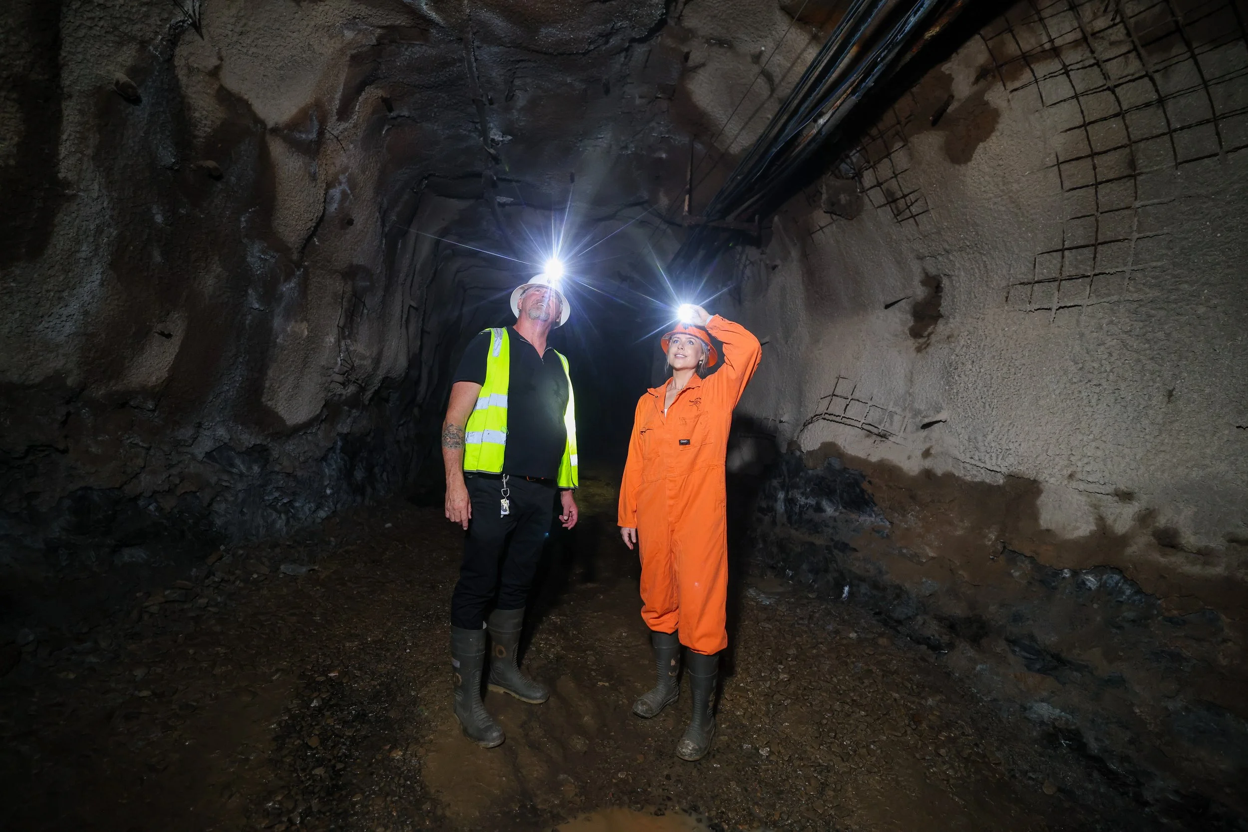 A mine tour guide with a guest inspecting a tunnel with headlamps within the Hard Times Mine. They are wearing safety gear including hard hats, reflective vests, and waterproof boots. The tunnel has exposed reinforcement bars and electrical conduits.