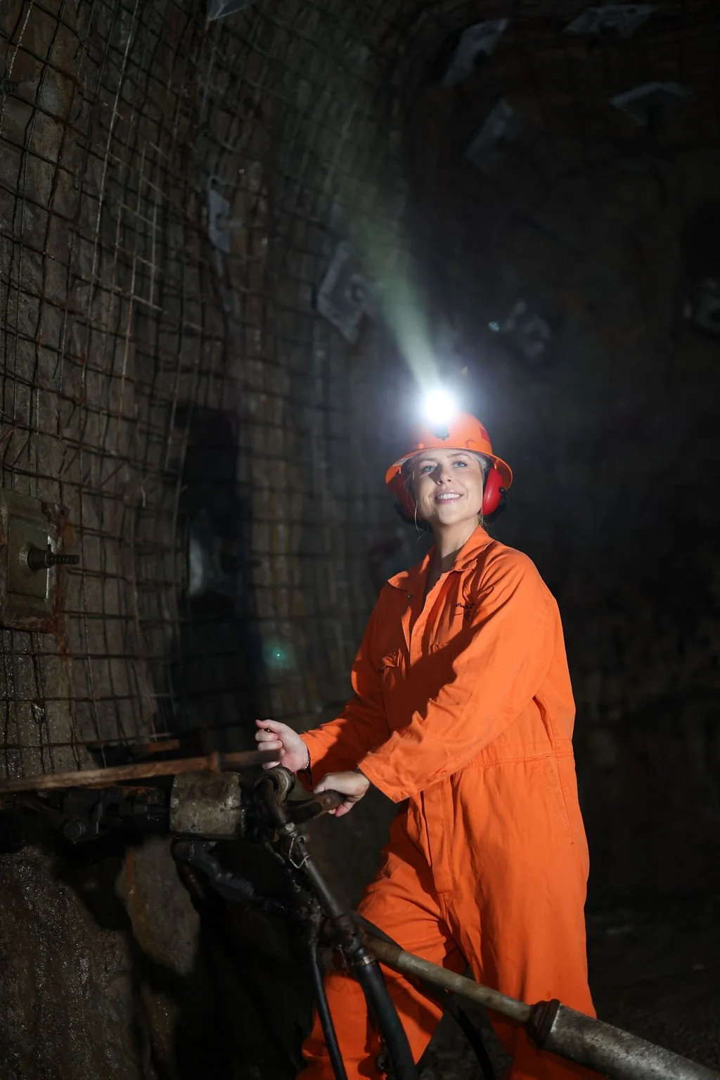 A woman in orange mining gear, including a hard hat with a headlamp and ear protection, operating machinery inside a tunnel with steel reinforcement.