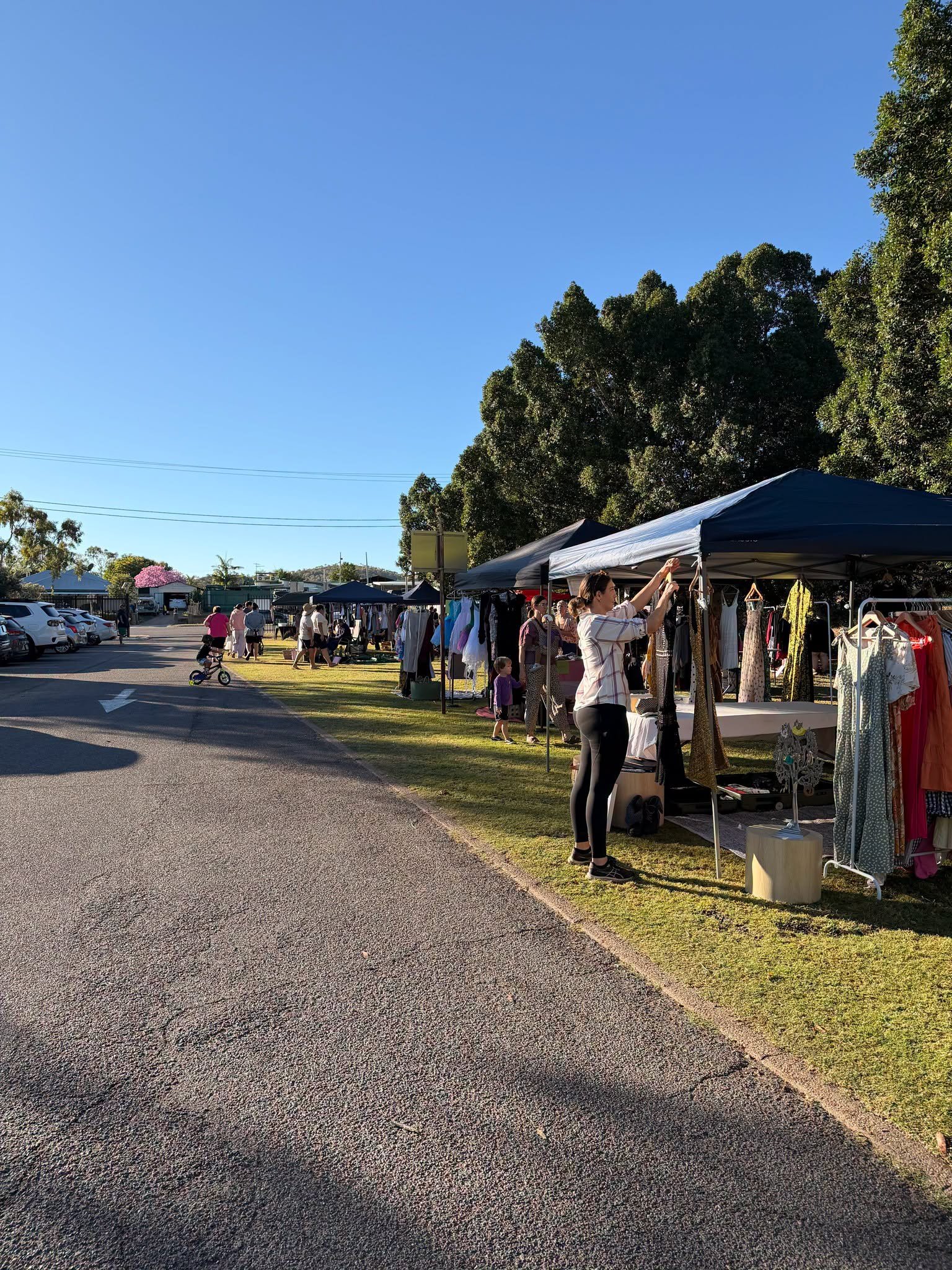Outdoor market set up with clothing stalls under tents, people browsing, in a parking lot area with trees and a clear blue sky.