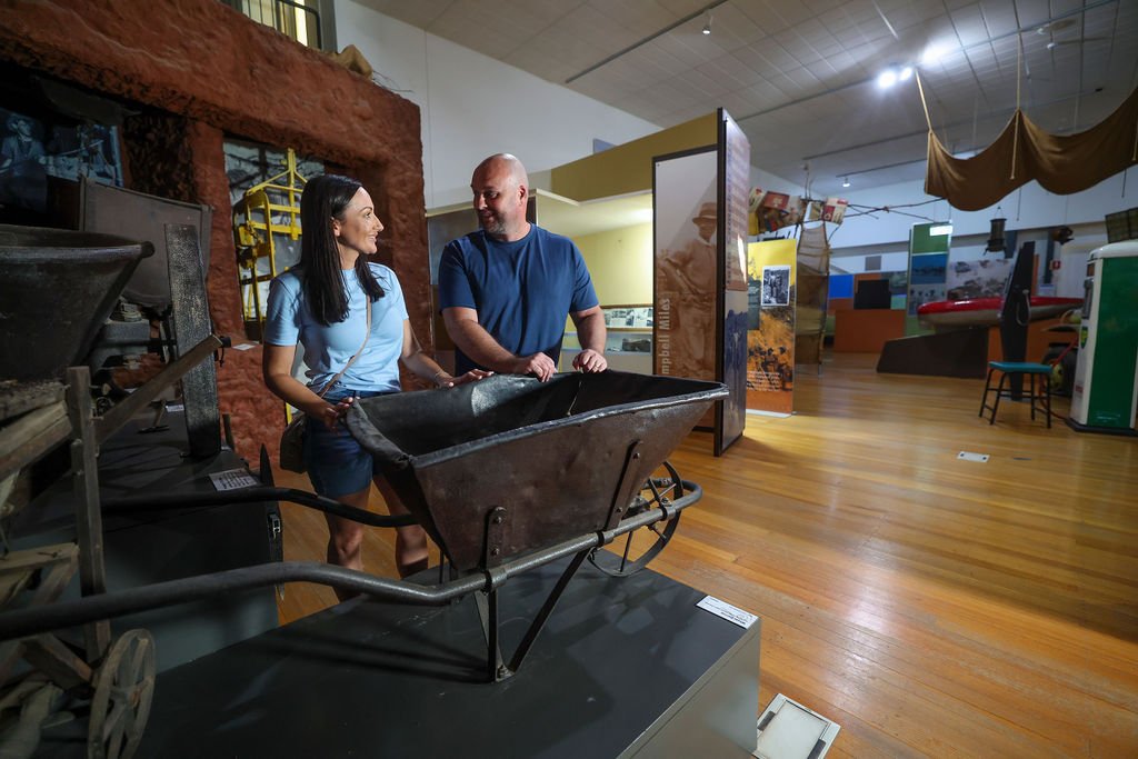 Two people smiling and talking in a museum exhibit, with an old wheelbarrow in front of them and display panels in the background.