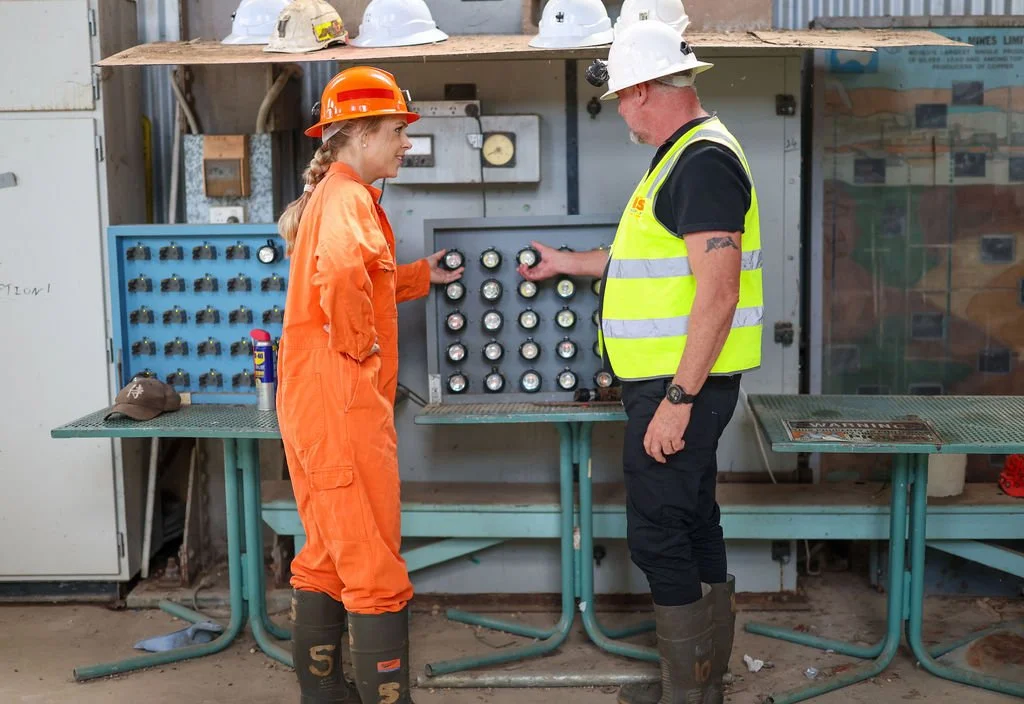 A woman in orange safety gear and a white hard hat holding a gauge, talking to an older man in a high-visibility vest and white hard hat checking gauges on a panel in an industrial or electrical setting.