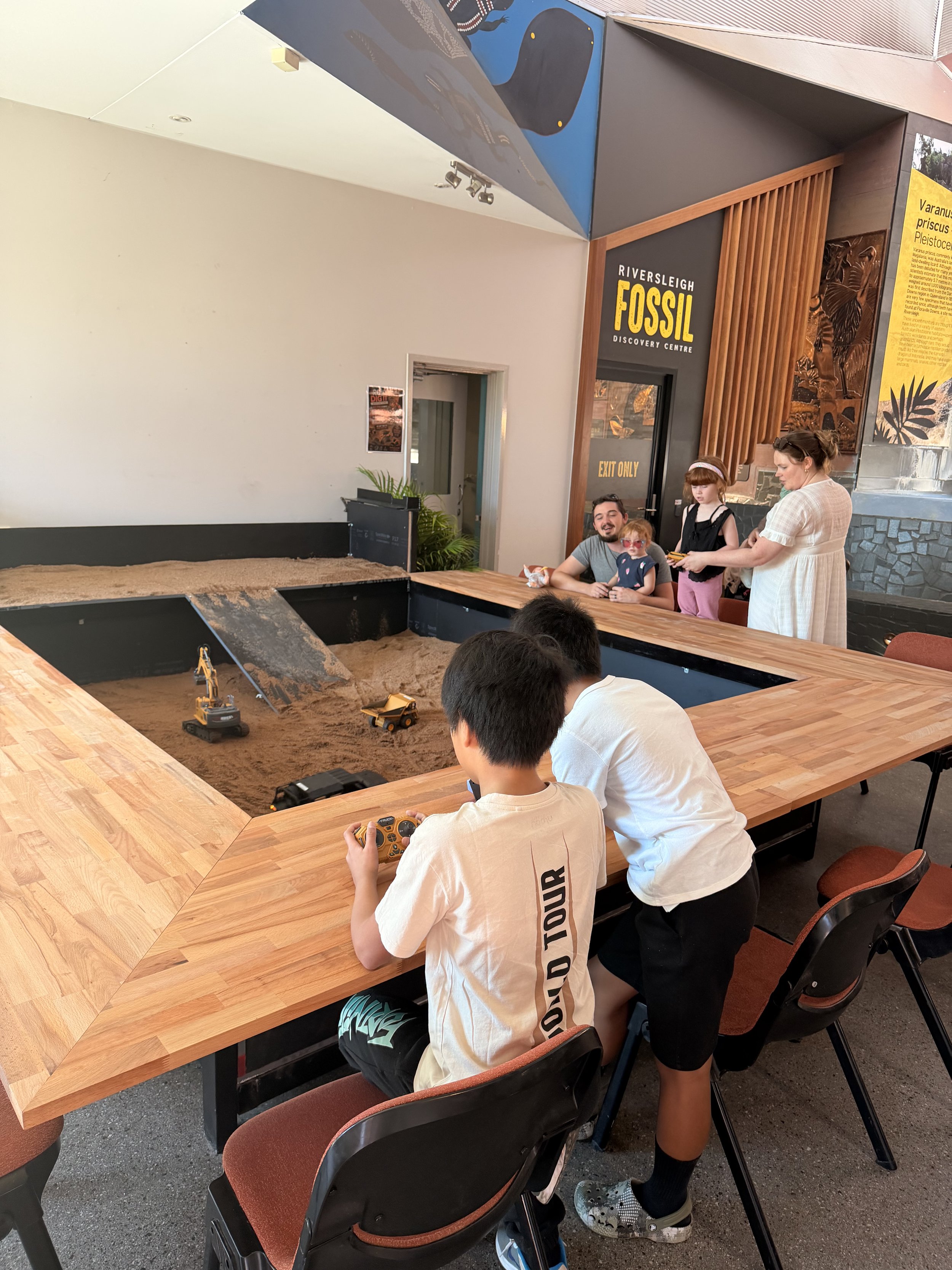 Children playing with toy excavators in a sandpit at the Outback Cafe, with an adult supervising. The scene includes a sand area, wooden tables, and educational display boards.