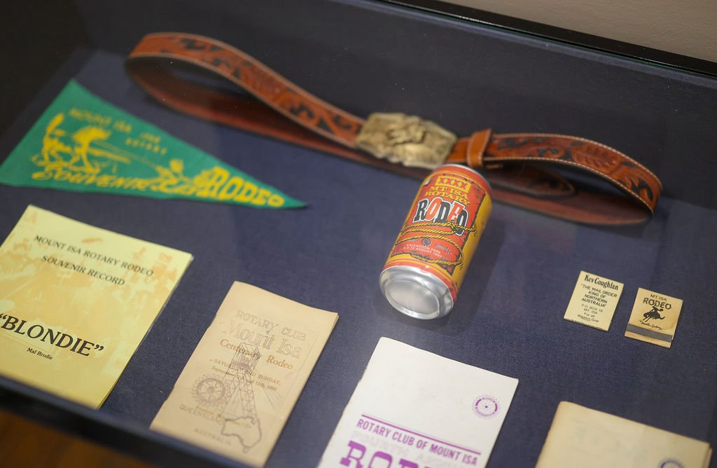 Display case in the Isa Rodeo Hall Of Fame containing vintage items including a Rode beer can, a leather wristband, a green and yellow pennant, several small paper and cardboard documents related to Rotary Club and Rodeo, and other memorabilia.