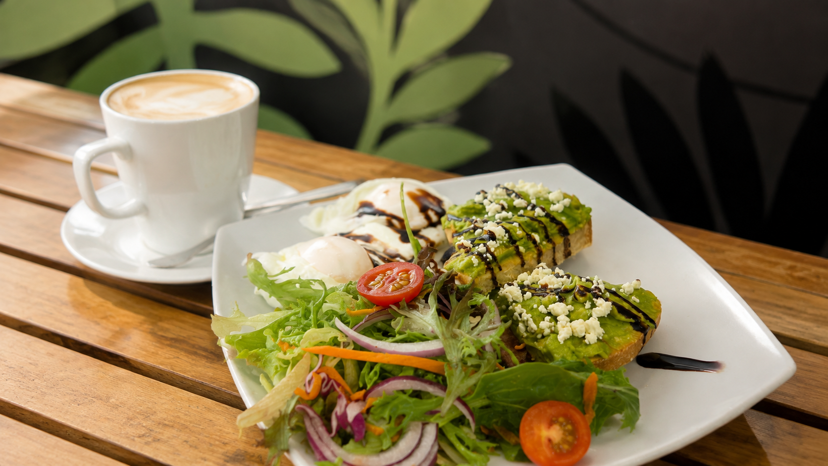 A white plate with avocado toast topped with cheese crumbles and drizzled with balsamic glaze, a side salad with cherry tomatoes, mixed greens, and sliced red onions, along with two poached eggs with balsamic glaze, next to a cup of coffee with foam on top, on a wooden table.