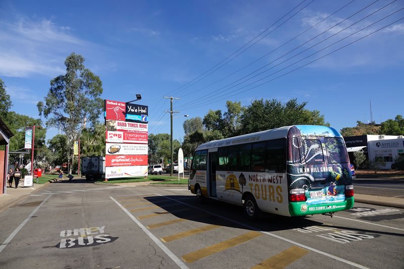 North West Tours bus parked in the designated bus zone at Outback At Isa, with trees in the background under a clear blue sky.