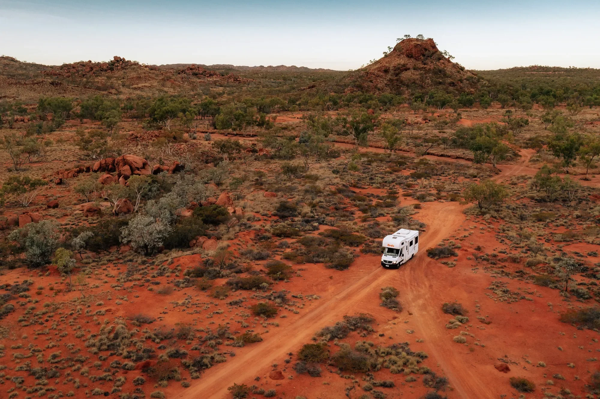 A white camper van traveling on a dirt road through a desert landscape with red rocks and sparse vegetation, under a clear sky.