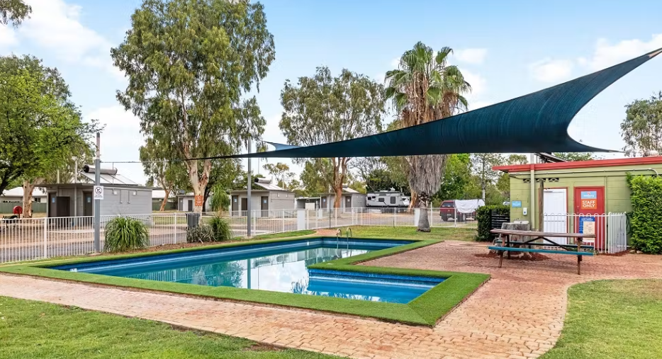 Public swimming pool with a shade sail overhead, surrounded by brick paving, green lawns, and trees, with a small building labeled 'Staff Only' nearby.