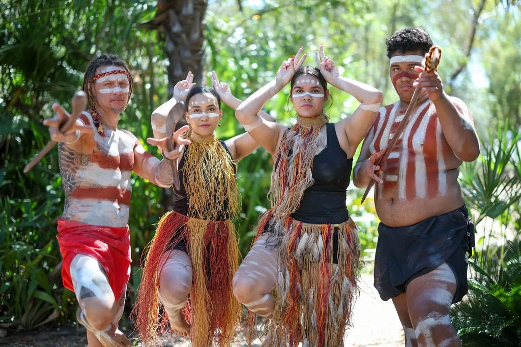 Four indigenous people dressed in traditional attire, painted with white patterns, posing outdoors with greenery in the background, holding spears and making gestures.