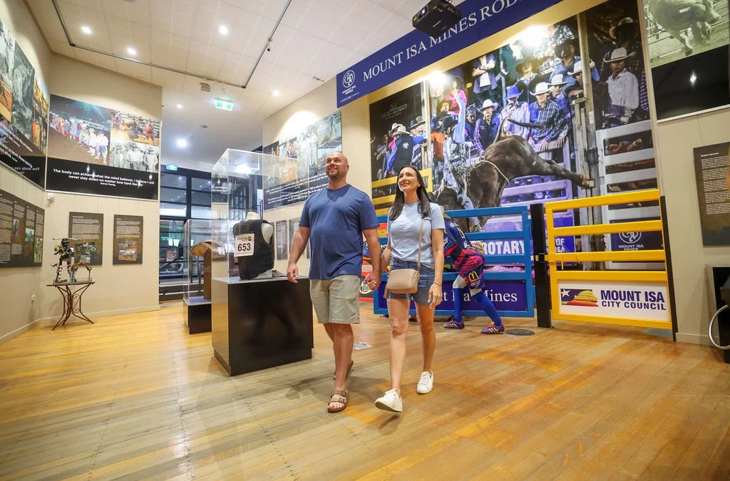 A couple holding hands walking through the Isa Rodeo Hall Of Fame exhibit with photos, displays, and a large mural of children and a horse behind a fence.