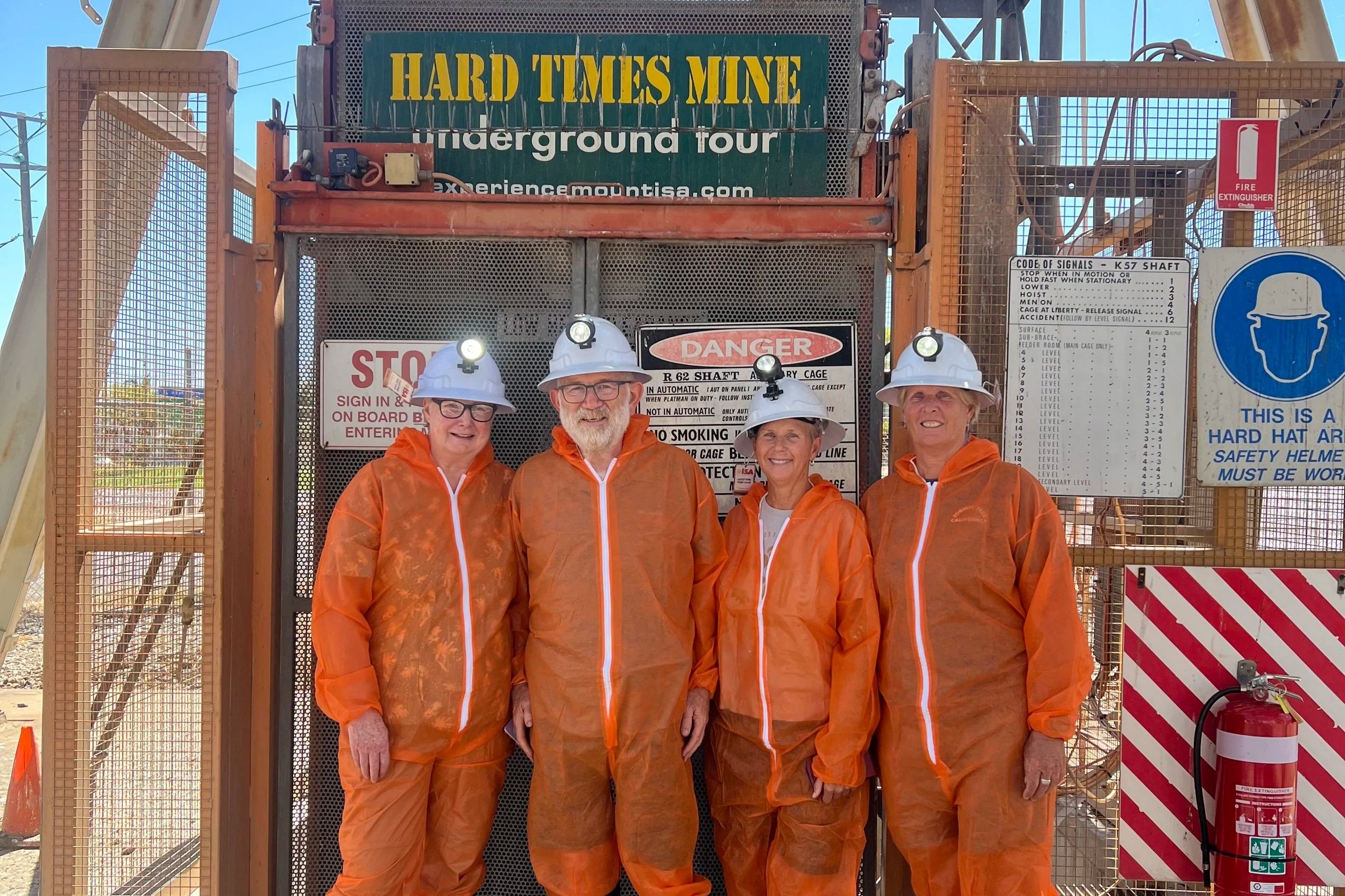 Four people dressed in orange safety gear and hard hats standing in front of a mine entrance sign that reads 'Hard Times Mine underground tour'.