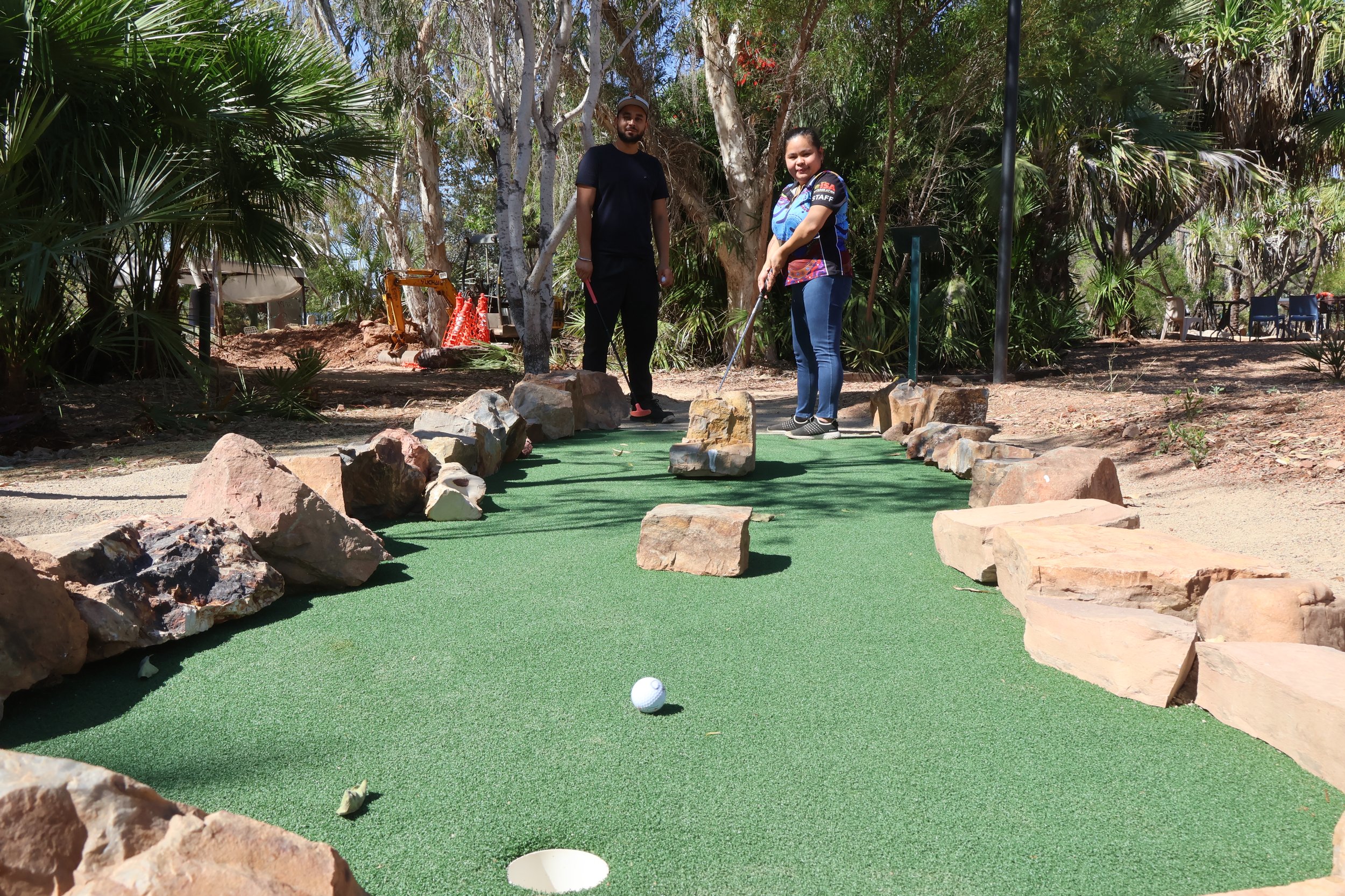 Two people playing mini golf outdoors at Outback At Isa on a green artificial turf course surrounded by trees and rocks, with a small white golf ball in the foreground.