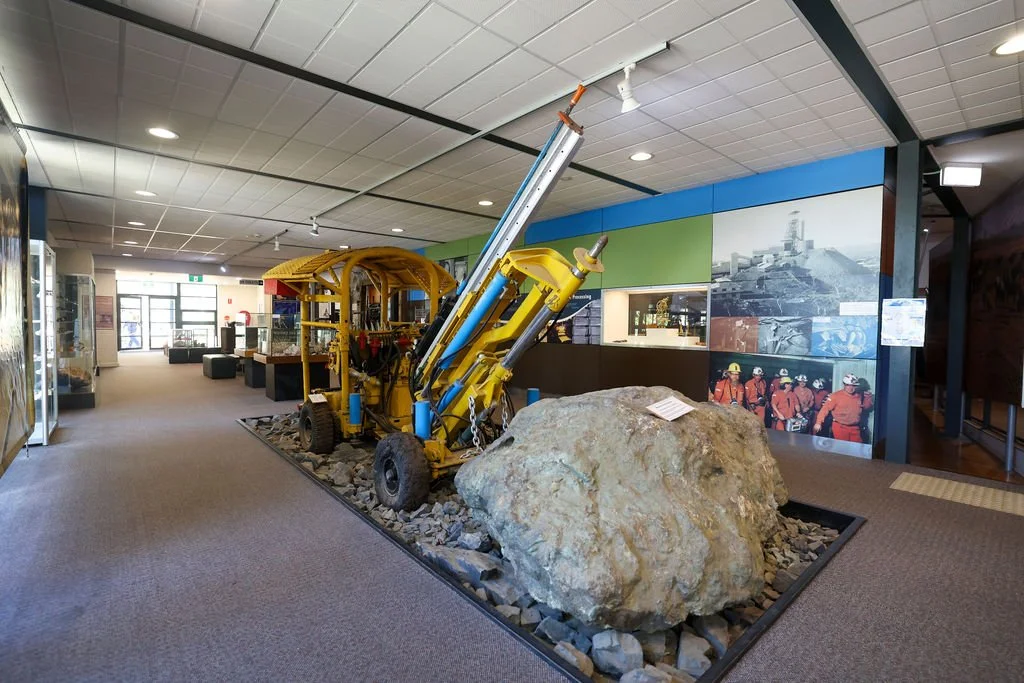 Museum exhibit featuring a large yellow underground mining machine and a big rock, with historical photos of miners on the wall in the background.