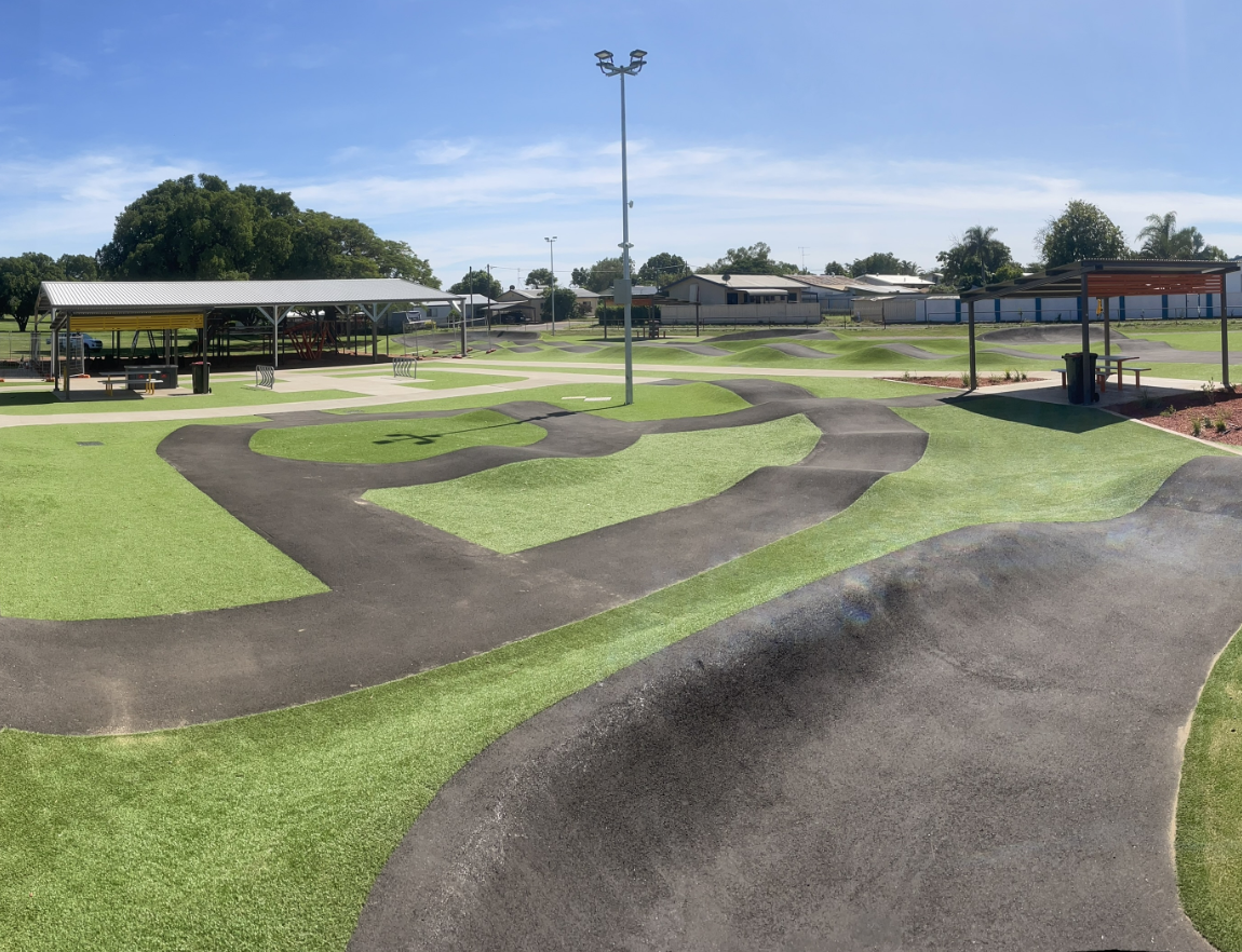 Empty outdoor pump track in Mount Isa with winding asphalt ramps and green grassy areas under a blue sky.