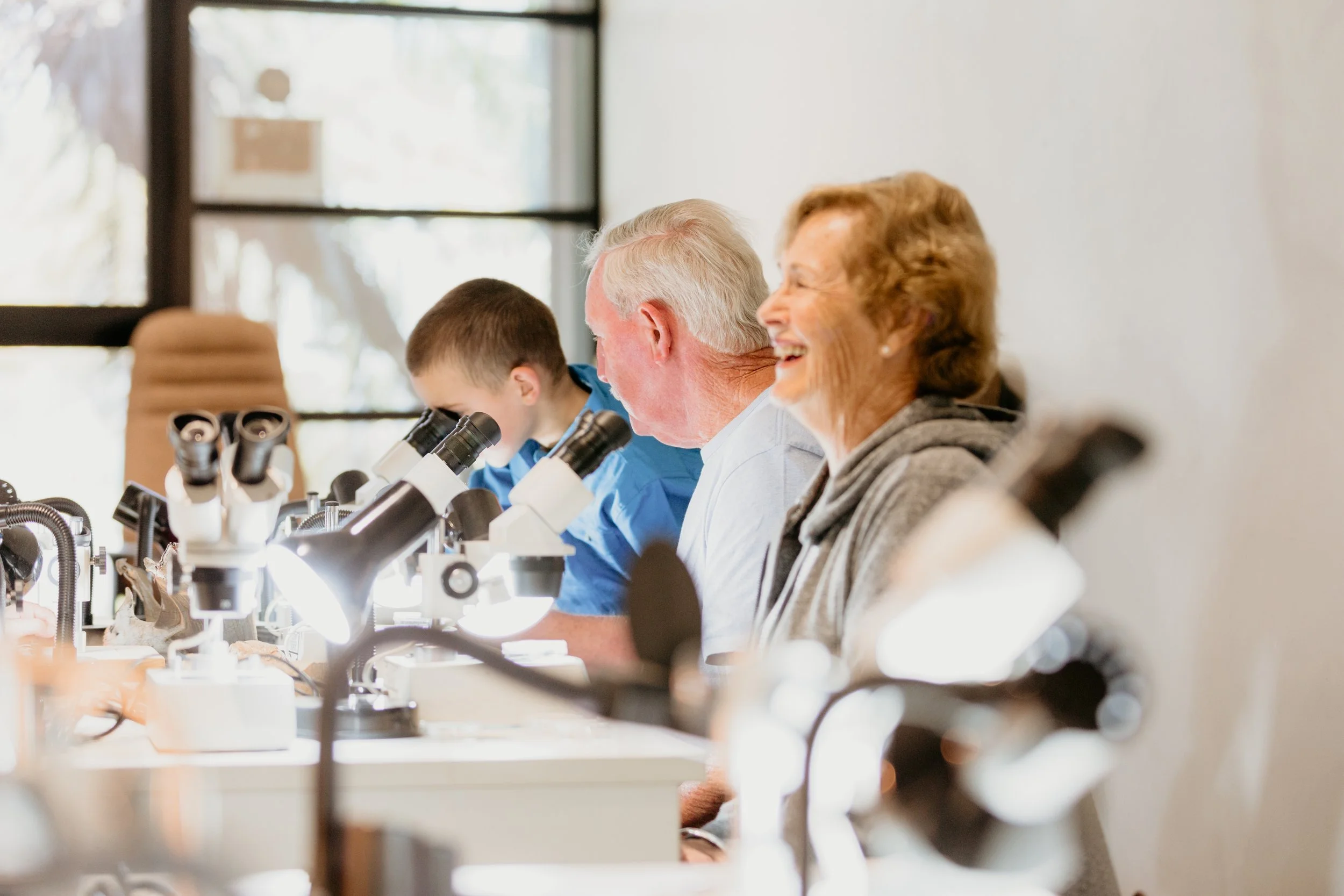 A diverse group of people, including elderly and young children, working with microscopes in a bright lab with large windows, engaging in scientific research or learning.