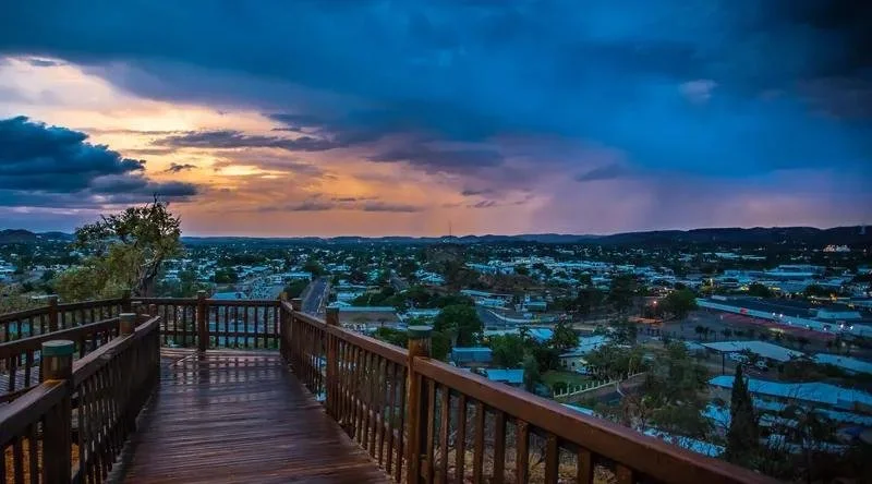 View of a city at sunset from a wooden observation deck with a railing, cloudy sky with hints of orange and purple