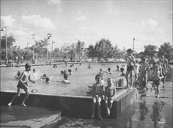 Historical Photo of Mount Isa with children and adults at a swimming pool, some swimming, others sitting or standing on the pool deck, with trees and utility poles in the background.