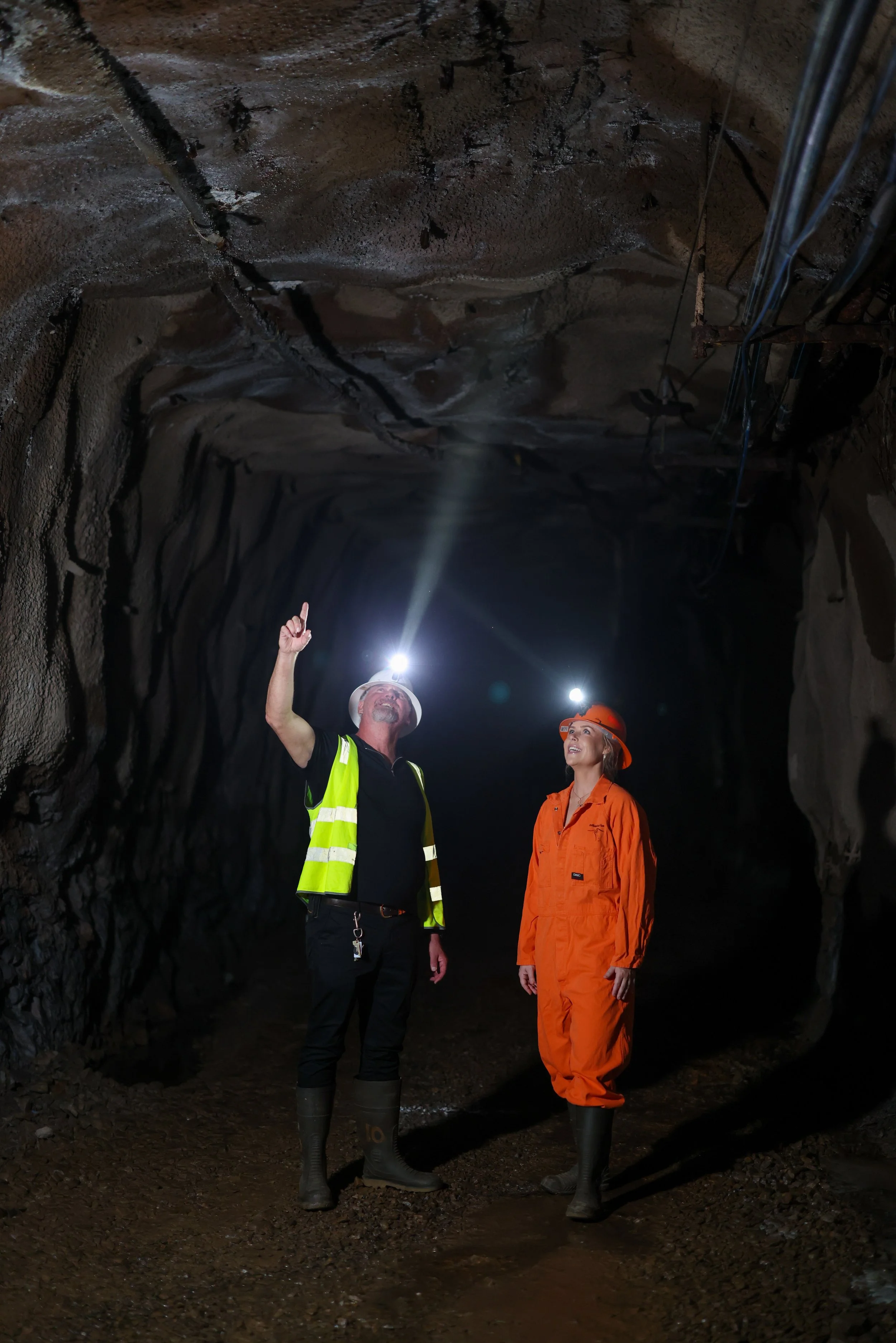 Two miners in an underground tunnel, one in a yellow safety vest and the other in orange coveralls, both wearing rubber boots and helmets with lights, looking up and pointing at the ceiling.