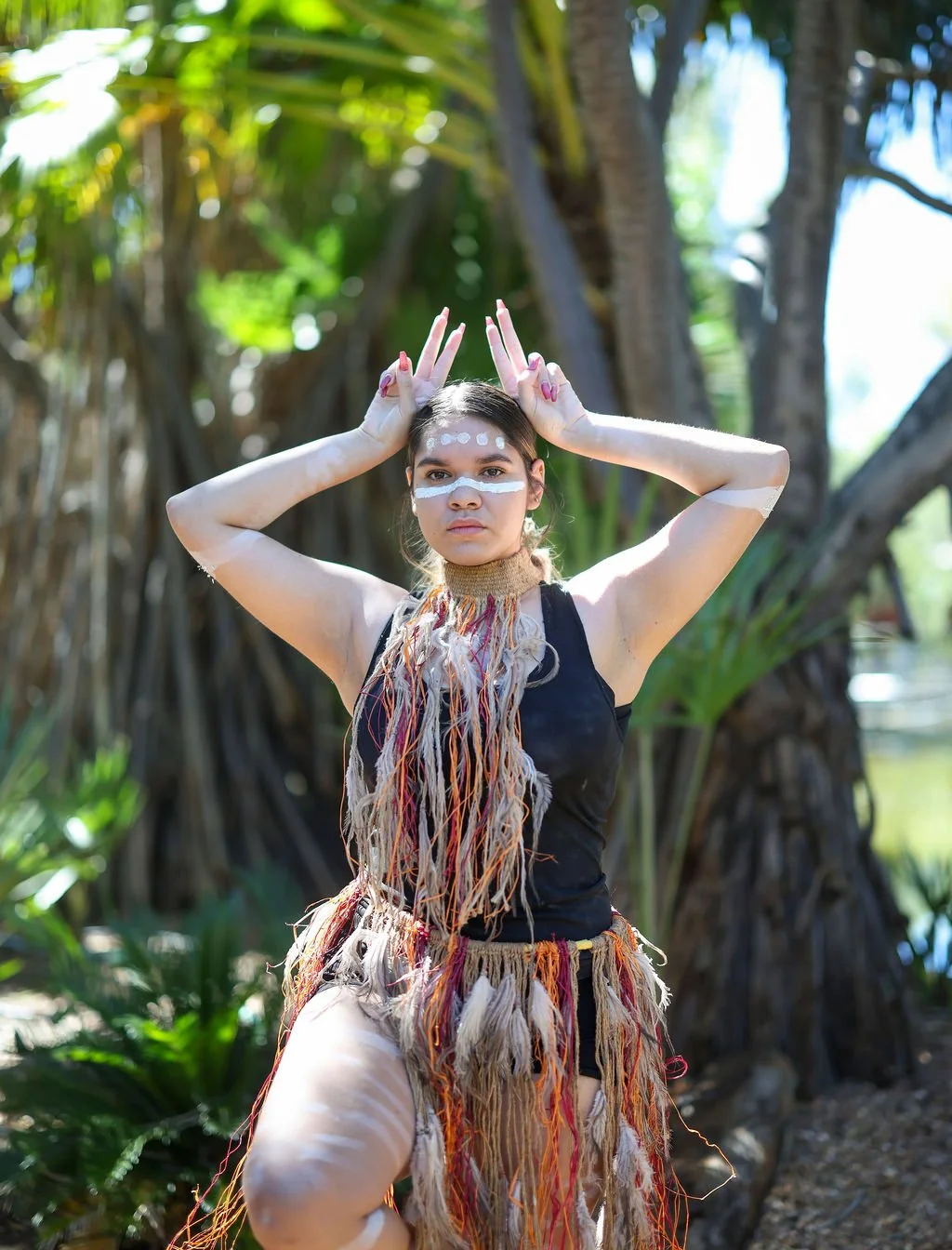 A woman with tribal-inspired face paint and feathered accessories poses outdoors, making a gesture with her hands resembling bunny ears, in front of lush greenery and trees.