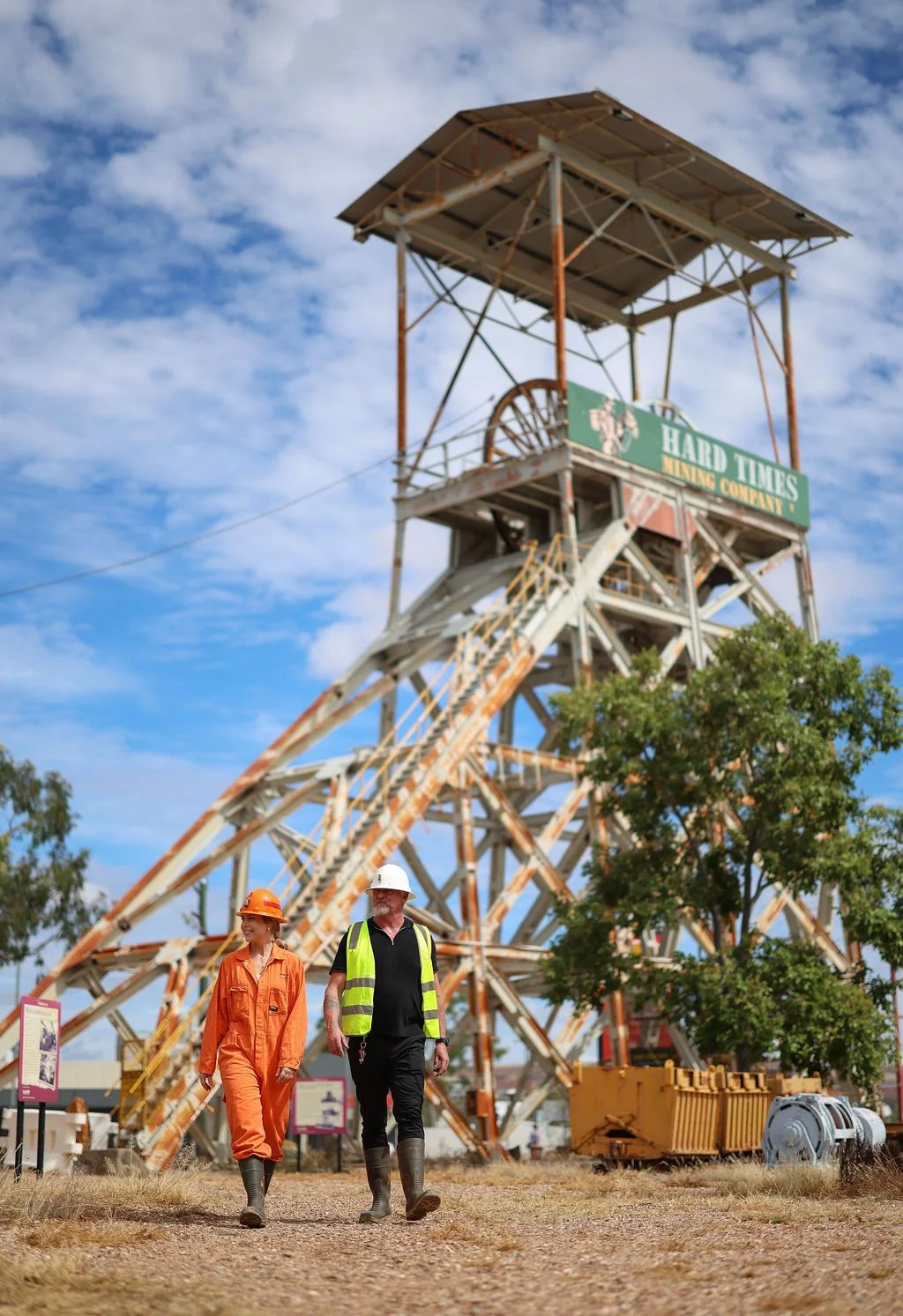 Two people dressed in Safety gear, a woman in an orange jumpsuit and hard hat, and a man in black clothing with a neon yellow safety vest and hard hat, walking in front of an old, rusty mining headframe with a sign that reads ‘HARD TIMES MINING COMPANY’ under a blue sky with scattered clouds.
