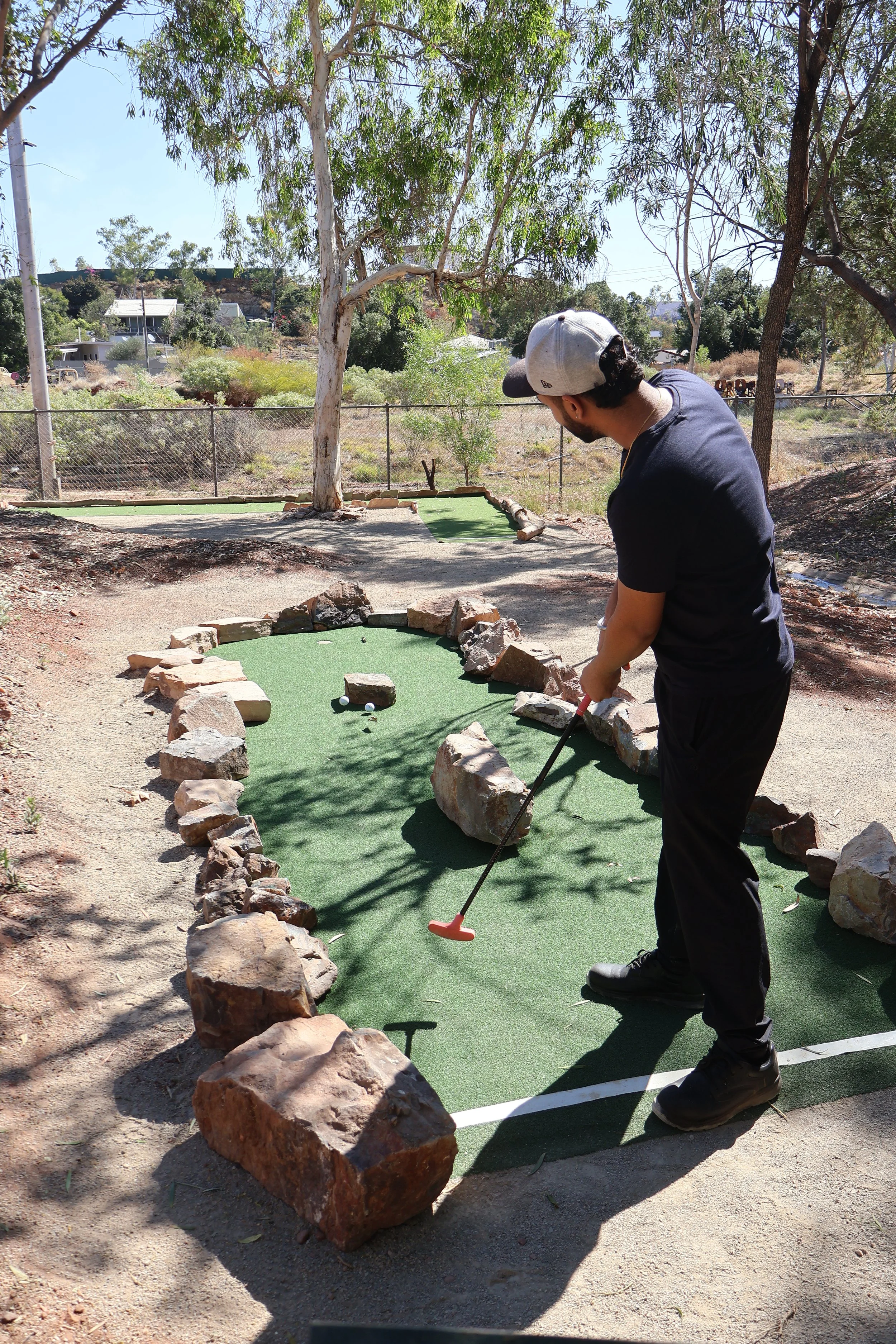A man wearing a gray cap and black sunglasses is playing outback mini golf on a green course surrounded by rocks. He is holding a golf club and preparing to hit a golf ball. There are trees and a fence in the background.
