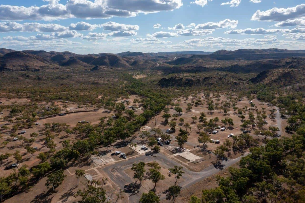An aerial view of a dry, mountainous landscape with sparse trees and a parking area with cars in the foreground, under a partly cloudy sky.