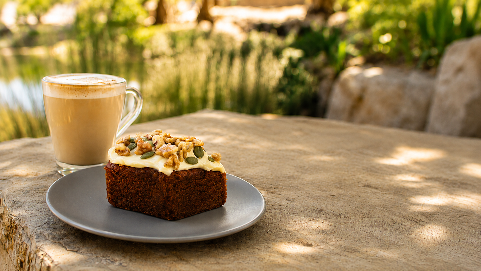 A plate with a piece of cake topped with white frosting, nuts, and seeds, a cup of coffee, on a stone table in the Outback At Isa Outback Park with a pond and green foliage in the background.