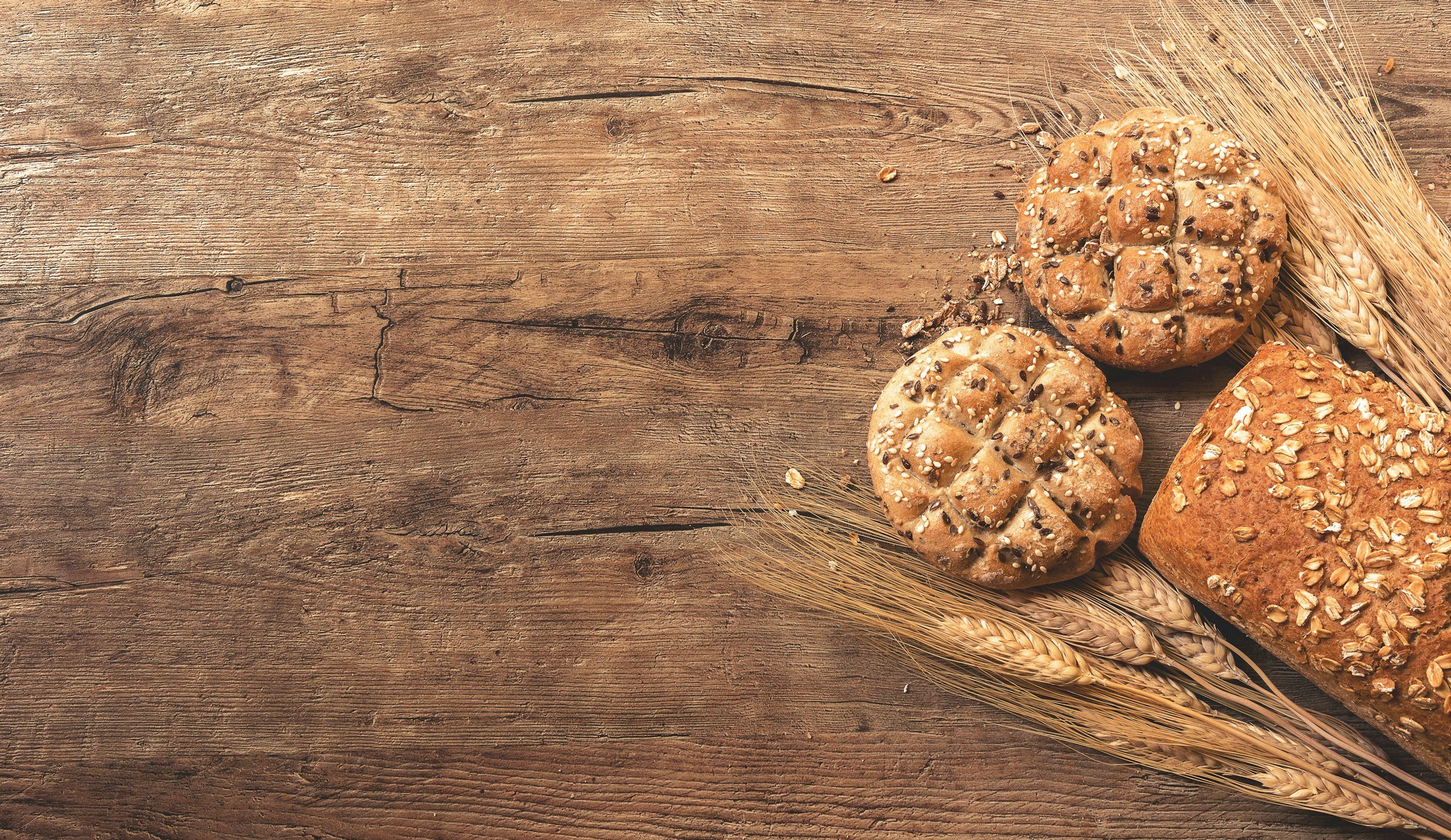 Several types of bread with oats and seeds on a rustic wooden surface, with wheat stalks beside them.