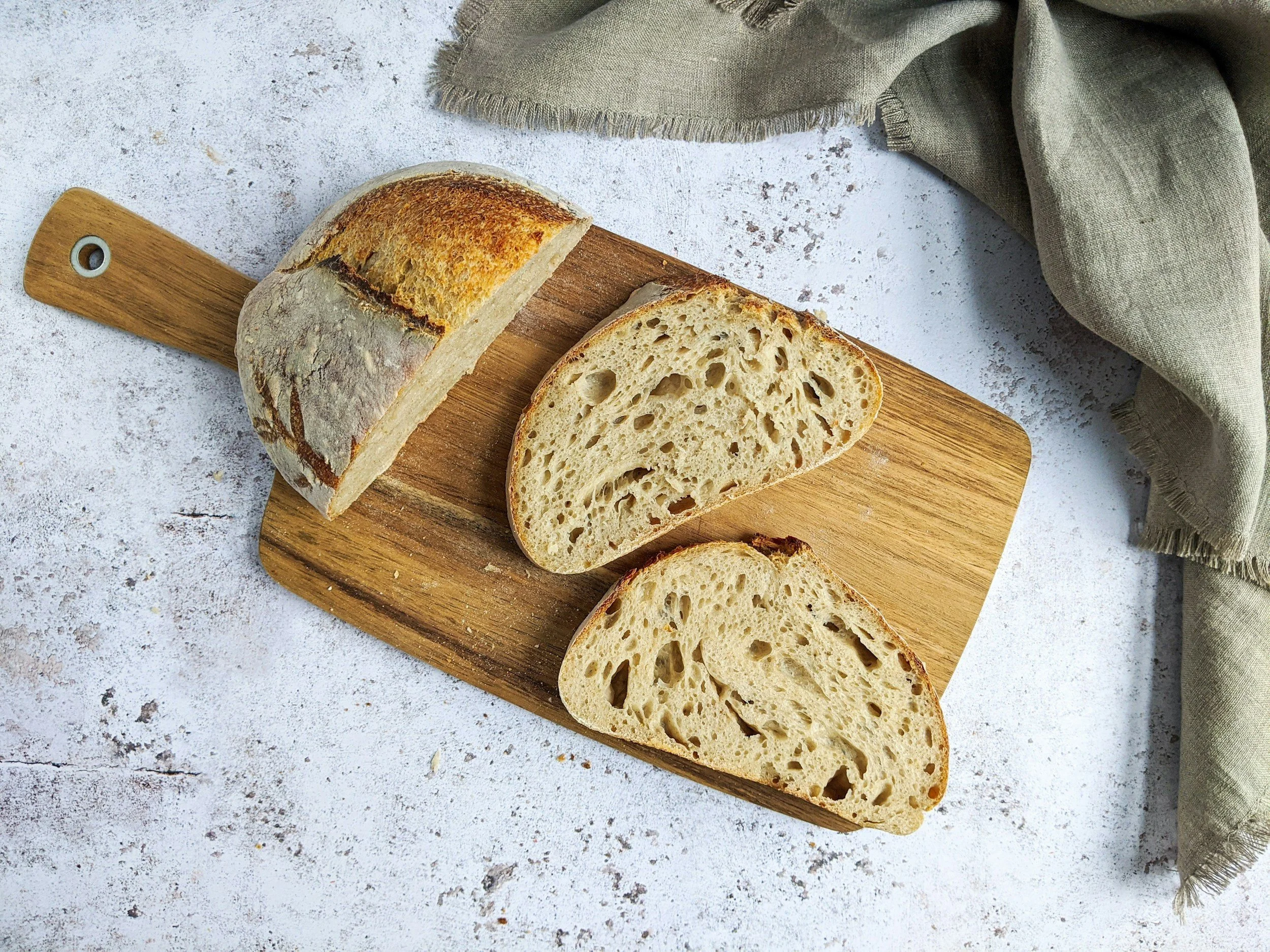 Sliced loaf of sourdough bread on a wooden cutting board, with a linen cloth nearby, on a textured white surface.