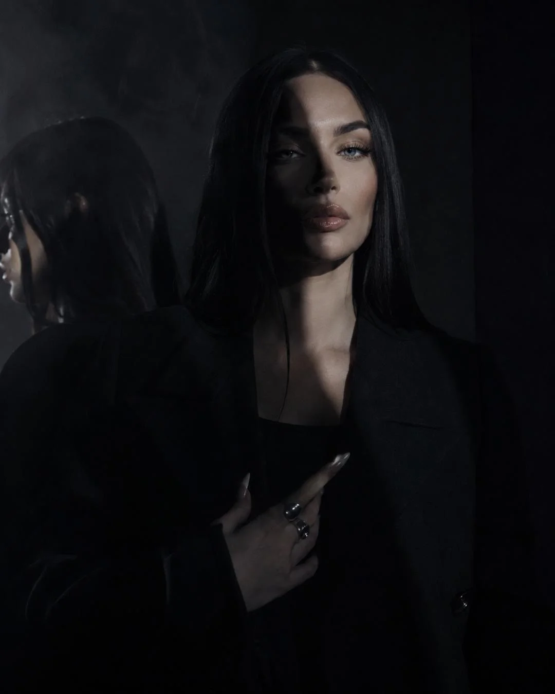 Dark cinematic portrait of SABLE, a woman with long black hair wearing a black coat, standing in low shadow beside a mirror with soft smoke and moody lighting.