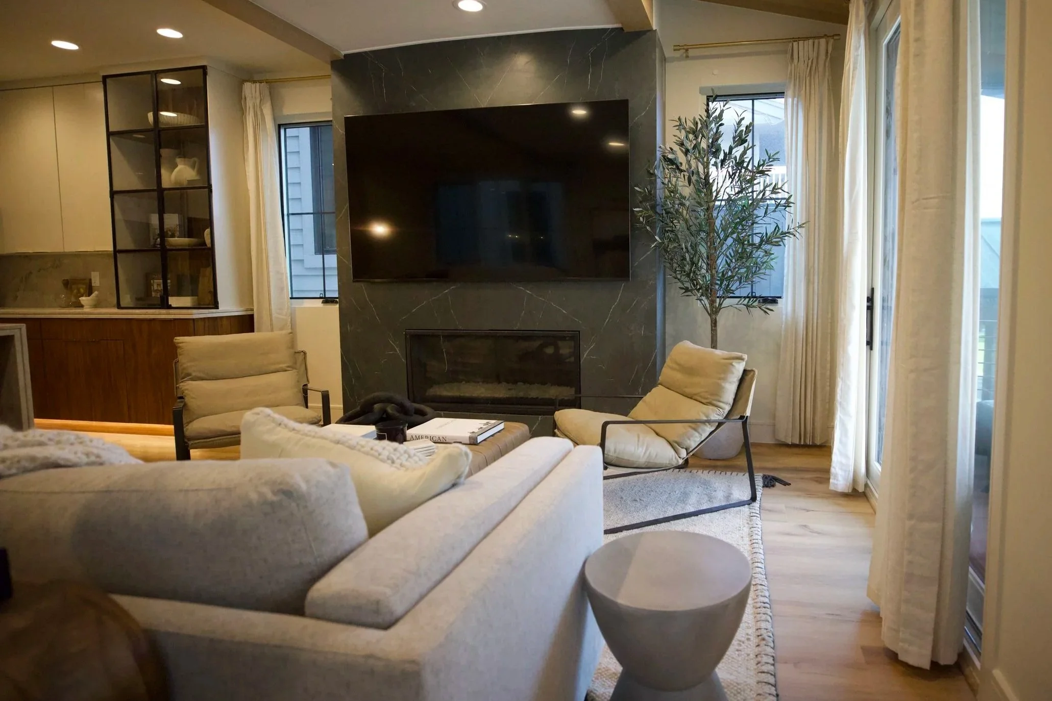 Living room with beige sofas, a black marble fireplace, a large flat-screen TV on a black marble wall, a tall green potted plant, and large windows with cream curtains.