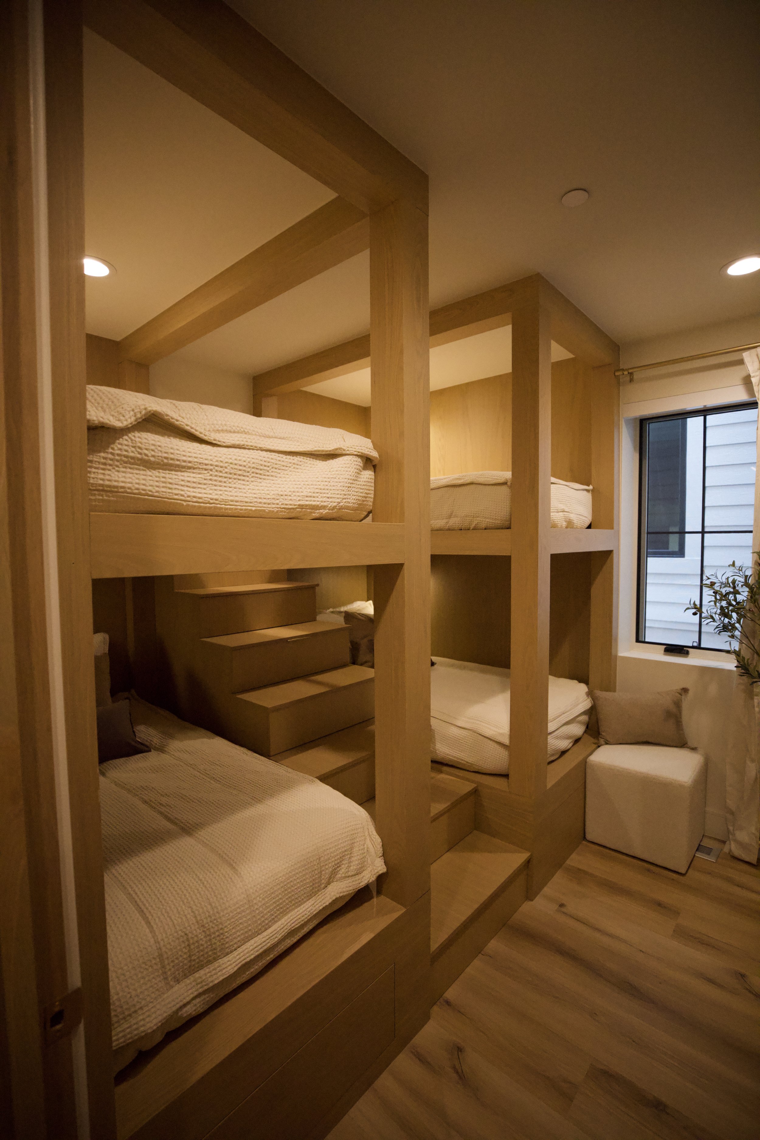 A bedroom with wooden bunk beds forming a unique structure with two beds on each level. The beds are made with beige bedding, and there is a small set of stairs leading up to the top bunk. A large window with a black frame and a white curtain is on one wall, allowing natural light into the room. There is a beige cushion on a small bench at the foot of the bottom bunk, and a plant is visible outside the window.
