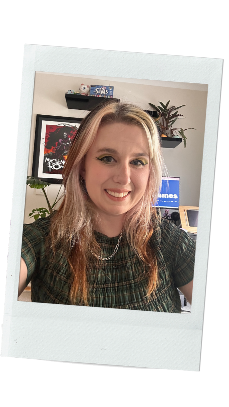 A woman with long, wavy, light brown hair and makeup, smiling in a selfie. She is wearing a green plaid top and a silver chain necklace. In the background, there are plants, a framed poster, a computer monitor, and shelves with game boxes and toys.